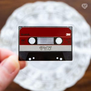 Op de afbeelding: Een doorzichtige sticker met een retro cassetteband ontwerp. De tape heeft een rode bovenkant, een zilveren midden en een zwarte onderkant. De datum "9/4/73" is handgeschreven op de tape. Het merk "Scotch" is zichtbaar.