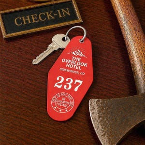 Könnte beinhalten: Ein roter, tropfenförmiger Hotel-Schlüsselanhänger aus Kunststoff mit einem silbernen Metallschlüssel. Der Anhänger trägt weißen Text mit der Aufschrift THE OVERLOOK HOTEL, SIDEWINDER, CO, die Nummer 237 und ein kreisförmiges Logo mit DROP IN ANY MAIL BOX WE GUARANTEE POSTAGE.
