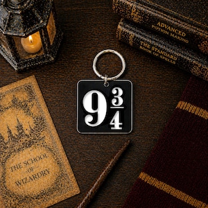 May include: A black square keychain with the number 9 3/4 in white. The keychain is on a silver ring. The image also includes a lit candle, books, a wand, and a scarf.