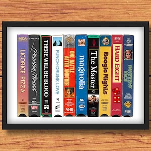 May include: A framed print featuring a collection of VHS tapes with movie titles like "Licorice Pizza," "Phantom Thread," and "Magnolia." The tapes are arranged vertically, showcasing their colorful spines against a white background.