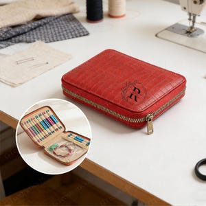 Peut inclure: Un étui à aiguilles à tricoter rouge et rectangulaire avec un motif texturé en forme de crocodile. L'étui a une fermeture éclair dorée et un monogramme avec la lettre "R" dans une couronne. L'étui ouvert révèle diverses aiguilles à tricoter et accessoires.