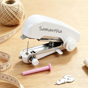 May include: A white handheld sewing machine featuring the name Samantha printed in black on the side. The device includes a metal needle, a white tension dial, a spool holder, and a small pink plastic bobbin pin positioned in front of it.