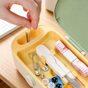 May include: A yellow and cream-colored sewing kit with a green lid. The kit contains needles, safety pins, buttons, a measuring tape, and small white scissors. The kit is open, and a hand is reaching in to grab a needle.