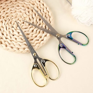 Könnte beinhalten: Zwei Scherenpaare mit Metallklingen und Griffen. Ein Paar hat goldfarbene Griffe mit dem Text "Aqualin", und das andere hat schillernde Griffe mit dem Text "Norman". Die Scheren sind für Bastelarbeiten oder Nähen konzipiert.