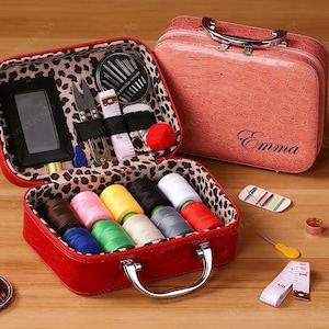May include: Red sewing kit with a leopard print lining, containing spools of thread, needles, scissors, and a measuring tape. A matching pink case with the name "Emma" is also present. Additional sewing notions are scattered around.