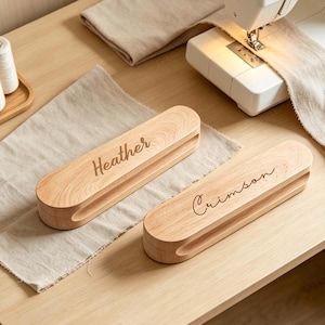 May include: Two elongated, light-coloured wooden clapper tools with rounded ends. One tool is engraved with the name Heather in a script font, and the other is engraved with the name Crimson in a script font. Both items feature a central slot.