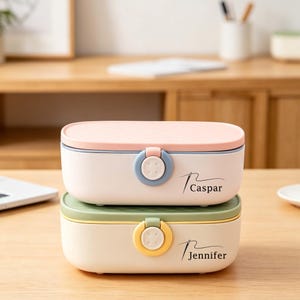May include: Two stacked, rectangular storage boxes with pastel pink and green lids. The top box has the name "Caspar" printed on it, and the bottom box has the name "Jennifer". Each box has a circular clasp.