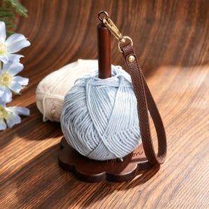 May include: A light blue yarn ball sits on a dark brown wooden stand with a flower-shaped base. A brown faux leather wrist strap with a gold metal lobster clasp is attached to the top of the wooden post. A cream yarn ball is visible behind.