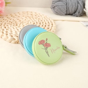 May include: Three pastel-coloured round measuring tapes. The green tape features a floral design and the name "Nicola". The tapes are stacked, with grey, blue, and green visible. Each tape has a small green pull tab. Metric measurements.
