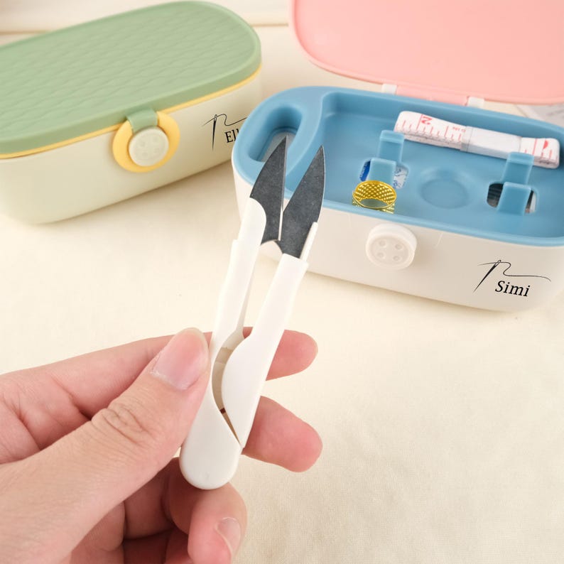 May include: White handled thread snips with black blades are held in front of a blue and white sewing kit. The kit contains a measuring tape and a gold thimble. A green and pink sewing kit are in the background. The text "Simi" is on the blue kit.