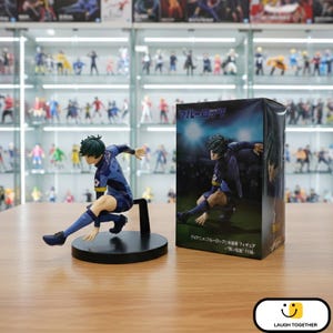 Może przedstawiać: Figurka piłkarza w niebieskim uniformie, w akcji, na czarnej podstawie. Obok pudełko z japońskim tekstem i wizerunkiem tego samego zawodnika. W tle gablota wypełniona innymi figurkami.