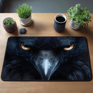 May include: Black rectangular mousepad featuring a close-up of a raven's face with intense golden eyes. The raven's feathers are detailed, creating a textured appearance. The mousepad is designed for computer use and adds a striking visual element to any desk setup.
