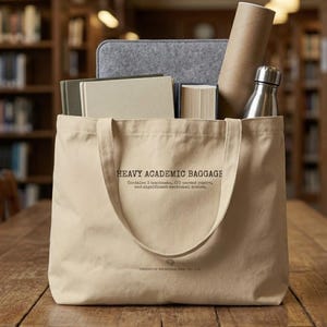 May include: A beige canvas tote bag containing books, a grey laptop sleeve, a brown paper tube, and a silver water bottle. The bag features the text "HEAVY ACADEMIC BAGGAGE".