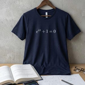 Op de afbeelding: Marineblauwe t-shirt met de wiskundige vergelijking "e^iπ + 1 = 0" in het wit. Het shirt hangt aan een houten hanger. Een leerboek, een rekenmachine en een bril liggen op een houten oppervlak.