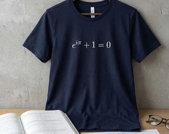 May include: Navy blue t-shirt with the mathematical equation "e^iπ + 1 = 0" printed in white. The shirt is hanging on a wooden hanger. A textbook, calculator, and eyeglasses are on a wooden surface.
