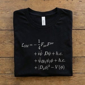 Peut inclure: T-shirt noir avec texte blanc affichant une équation de physique. Le vêtement est plié proprement sur une surface en bois. Le texte comprend des symboles mathématiques et des lettres grecques.
