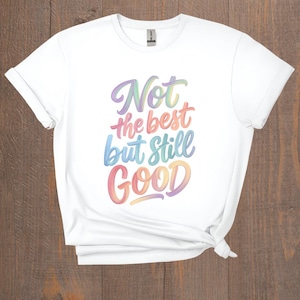 May include: White t-shirt with the phrase "Not the best but still Good" in colorful, cursive lettering. The shirt is made of a soft material and has a classic crew neck design. The shirt is knotted at the bottom.