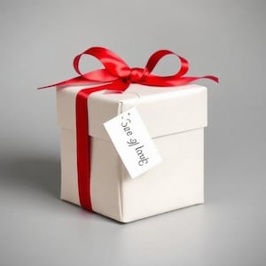 Puede incluir: Una caja de regalo blanca con una cinta y lazo de satén rojo. Una pequeña etiqueta blanca cuelga de la cinta con el texto "See if I laugh". La caja está sobre un fondo gris.