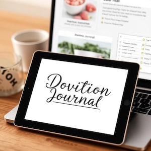May include: A black tablet displaying the text "Dovition Journal" in elegant script, resting on a laptop. A cup and a candle are in the background. The laptop screen shows a webpage with a journal theme.