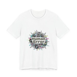Puede incluir: Camiseta blanca con un diseño gráfico que dice "Human Error". El diseño presenta elementos tecnológicos retro en gris, rosa y turquesa, dispuestos en un patrón circular. Una prenda de vestir casual.