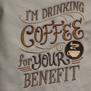 May include: A gray apron with brown embroidery that reads "I'm drinking coffee for your benefit". The embroidery includes a stylized coffee cup with steam.