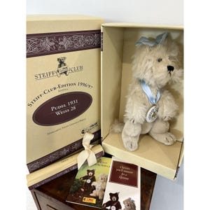May include: A cream-colored plush poodle toy with a light blue ribbon bow on its head and a matching blue ribbon around its neck holding a white Steiff Club medallion. The toy is presented in a beige box labeled Steiff Club Edition 1996/97.
