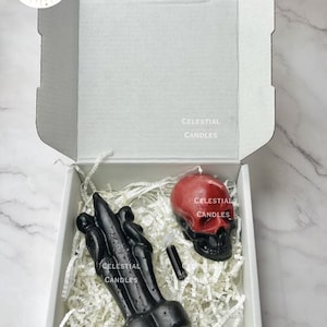 May include: A white box containing black candles and a red and black skull candle. The candles are surrounded by white paper filler. The box has the text "CELESTIAL CANDLES".