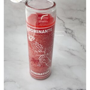 May include: A red wax candle in a clear glass cylinder container. The label features the text "DOMINANTE" and "DOMINATION" with a graphic of an eye, lightning bolts, and a hand. The brand name "CELESTIAL CANDLES" is printed on the glass.