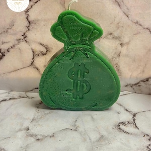 May include: A green candle shaped like a money bag, featuring a prominent dollar sign. The candle has a textured surface and a white wick. The background is a marble-patterned surface. The Celestial Candles logo is in the top left corner.