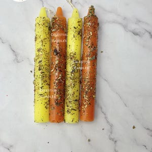 May include: Four cylindrical taper candles arranged vertically, alternating between bright yellow and orange colors. Each candle is coated with gold glitter and dried botanical herbs. The text Celestial Candles is printed on the candles.