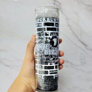 May include: A tall, cylindrical glass candle filled with black wax. The exterior features a white graphic print of a brick wall pattern with the text BLOCKBUSTER, ROMPE BLOQUE, OBEY ME, BUENA SUERTE, QUIEREM, LOVE, and VEN, along with a central moon icon.