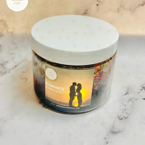 May include: A clear jar candle with a white lid. The label reads "Romance" with a silhouette of a couple kissing at sunset. The candle contains red and gold glitter. The brand name is Celestial Candles.