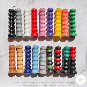 May include: A collection of colorful pillar candles in various shades, including black, gray, white, red, pink, purple, yellow, orange, green, and brown. Each candle has a unique stacked design. The candles are arranged on a white marble surface.