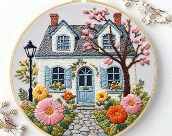 May include: Hand-stitched embroidery art of a charming cottage scene. The white house features a gray roof, blue shutters, and a stone pathway. Bright pink and orange flowers surround the house, with a flowering tree and a black lamppost.