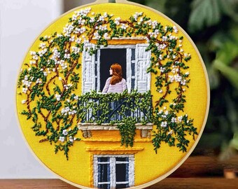 May include: A circular embroidery hoop featuring a yellow fabric background with a hand-stitched scene of a person with reddish hair standing at a window. The window is framed by white shutters and surrounded by dense green vines with white flowers.