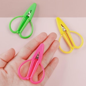 May include: Three small, colorful scissors in green, yellow, and pink. The scissors have rounded handles and a pointed blade. The scissors are made of plastic and are shown against a pink background.