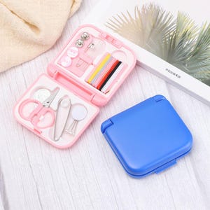 May include: A pink sewing kit with a variety of tools, including scissors, needles, buttons, and thread. The kit is open, revealing its contents, with a closed blue kit next to it. The kit is on a white wooden surface.
