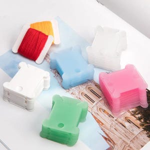 May include: A collection of colorful embroidery floss bobbins in white, pink, blue, and green. Two bobbins are filled with red and yellow thread. The bobbins are stacked on a printed surface, ideal for organizing embroidery floss.