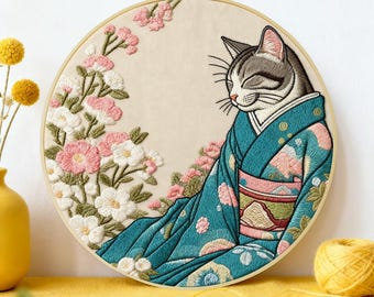 May include: A circular embroidery hoop containing a fabric panel with a stitched design of a grey and white cat wearing a teal patterned kimono. The cat is positioned next to a cluster of embroidered pink and white flowers on a cream background.