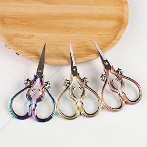 May include: Three pairs of metal craft scissors with pointed blades and ornate, curved handles. The left pair has a rainbow iridescent finish, the center pair is antique gold, and the right pair has a copper finish, all featuring small star accents.