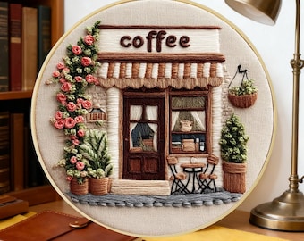 May include: A circular embroidery hoop featuring a hand-stitched scene of a coffee shop storefront. The design includes a brown door, a striped awning, pink climbing roses, a hanging basket, and a small table with chairs on a beige fabric.