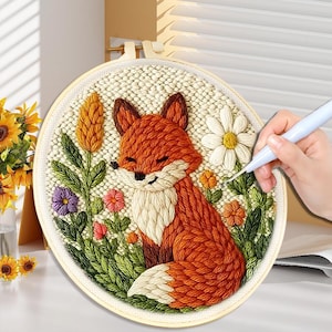 May include: A completed embroidery hoop featuring a smiling orange fox with white accents, surrounded by colorful flowers and greenery. The fox is the central focus, with a detailed, textured appearance. The embroidery is set in a cream-colored fabric within a wooden hoop.