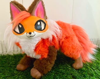 Puede incluir: Un juguete de peluche con forma de zorro que presenta una cara naranja pintada y lisa, grandes ojos negros, orejas con forro rosa y un mechón de pelo naranja. El cuerpo está cubierto de piel sintética naranja y blanca, con patas marrones.