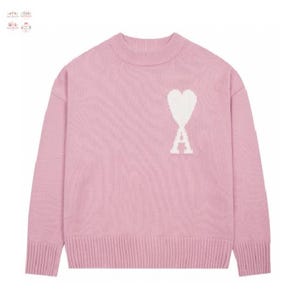 以下が含まれることがあります： 胸に白いハートと「A」のロゴが入った、明るいピンク色のセーター。クルーネックで、袖口と裾はリブ編み。柔らかく着心地の良いニットです。