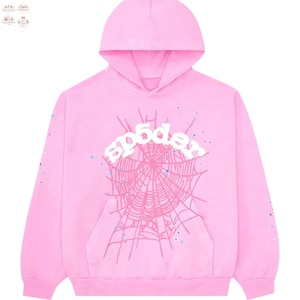 Puede incluir: Una sudadera con capucha rosa claro con capucha y bolsillo delantero. La parte delantera presenta un diseño de telaraña blanca con la palabra "sp5der" encima. Las mangas tienen pequeñas salpicaduras de pintura azules y blancas.