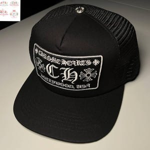 May include: Black trucker hat with a mesh back and a curved brim. The front panel features a white embroidered patch with the text "CHROME HEARTS" and "CH" with decorative floral accents. A silver emblem is on the top.