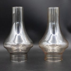 May include: Two clear glass lamp shades with a bulbous body and a cylindrical top. One shade has a slight amber tint. The shades are displayed against a dark background.