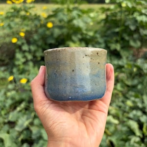 May include: A small, cylindrical ceramic cup featuring a textured surface with a gradient color scheme. The upper portion is a muted beige, transitioning into a deep blue at the base. The finish appears speckled and matte with visible clay texture.