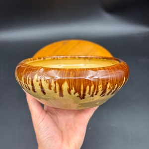 May include: A ceramic bowl with a wide rim and a shallow interior. The rim is a rich brown color with a dripped glaze effect, and the interior is a light yellow. The bowl is held in a hand.