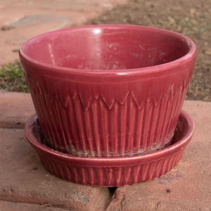 May include: A burgundy ceramic planter with a matching saucer. The planter has vertical grooves and a decorative zigzag pattern near the rim. The set is designed for indoor or outdoor use.